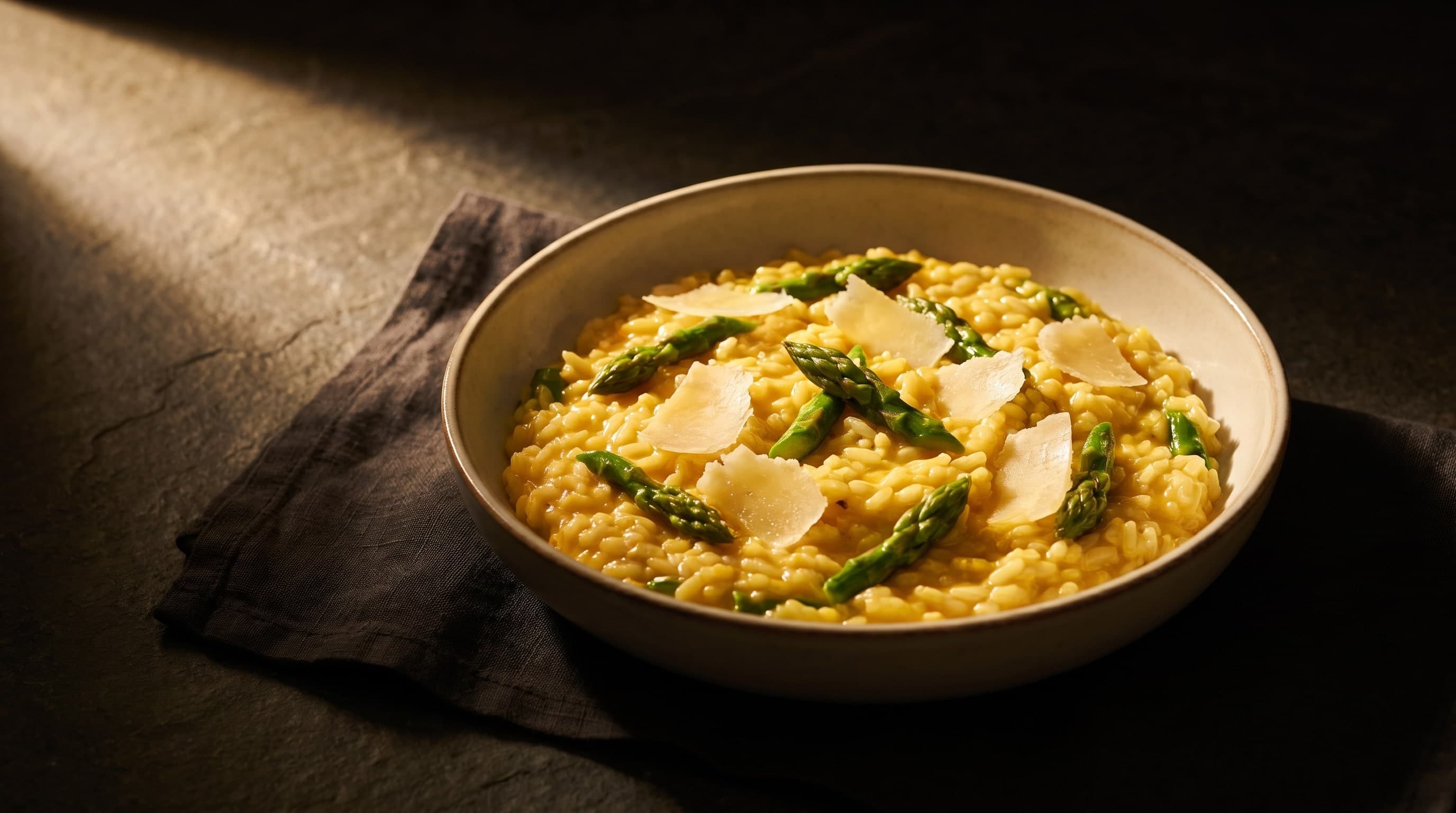 Risotto au safran, asperges vertes et copeaux de parmesan dans un bol clair sur pierre sombre